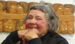 Photo of Mabel as an old woman. She has short grey hair and is smiling at the camera, leaning her chin on her hands. Behind her is a large carved wooden panel depicting many faces in two rows.